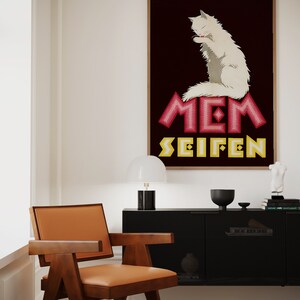 May include: A framed vintage advertisement featuring a white cat grooming itself, set against a dark burgundy background. The text "MEM SEIFEN" is displayed in pink and yellow. The room includes a wooden chair with an orange seat.