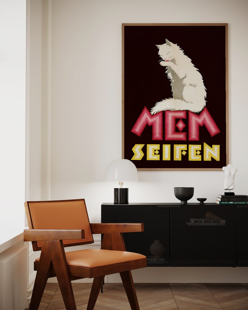 May include: A framed vintage advertisement featuring a white cat grooming itself, set against a dark burgundy background. The text "MEM SEIFEN" is displayed in pink and yellow. The room includes a wooden chair with an orange seat.