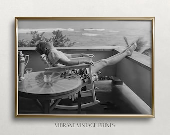May include: Black and white framed print of a person relaxing in a rocking chair on a balcony overlooking the ocean. A round table with a glass top and a cocktail shaker are in the foreground. The words "VIBRANT VINTAGE PRINTS" are at the bottom.