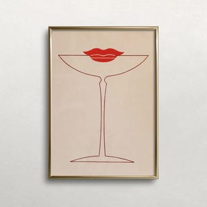 May include: A framed wall art print featuring a minimalist line drawing of a martini glass in dark red ink on a light beige background. A pair of solid red lips is positioned directly above the rim of the glass, creating a stylised graphic.