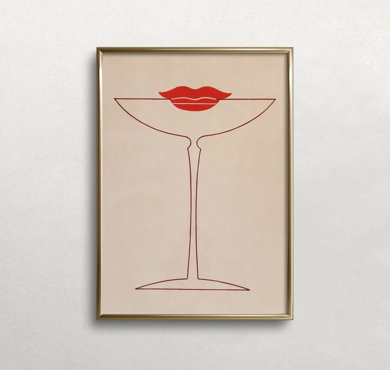 May include: A framed wall art print featuring a minimalist line drawing of a martini glass in dark red ink on a light beige background. A pair of solid red lips is positioned directly above the rim of the glass, creating a stylised graphic.