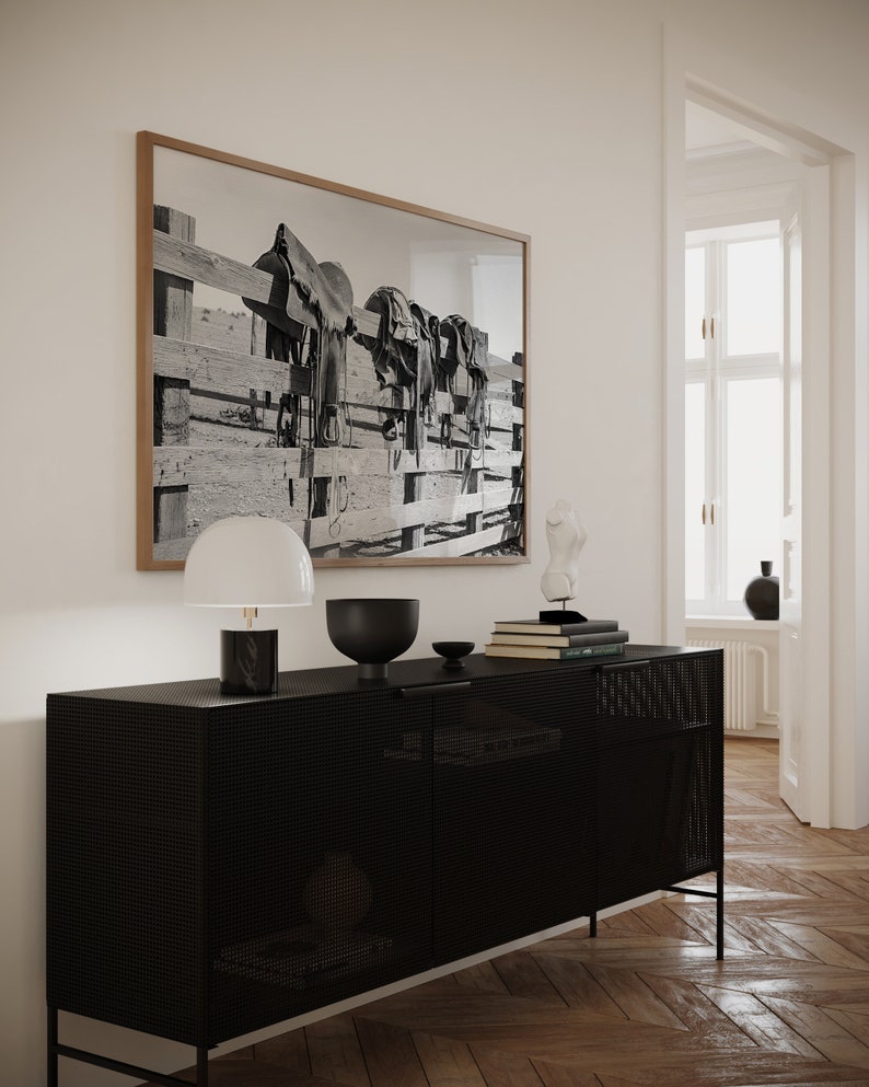 K&ouml;nnte beinhalten: Ein schwarzer Metallschrank mit einer Mesh-Front und zwei T&uuml;ren. Der Schrank steht auf einem Holzboden. Eine wei&szlig;e Lampe mit schwarzem Fu&szlig; steht auf dem Schrank, zusammen mit einer schwarzen Schale und einem Stapel B&uuml;cher. Ein gerahmtes Schwarzwei&szlig;foto von S&auml;tteln, die an einem Zaun h&auml;ngen, befindet sich an der Wand hinter dem Schrank.