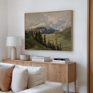 Op de afbeelding: Een rechthoekig landschapsschilderij in een lichthouten lijst met besneeuwde bergen, een groene vallei en een dennenbos. Het kunstwerk hangt aan een witte muur boven een lichthouten dressoir met boeken en een tafellamp.