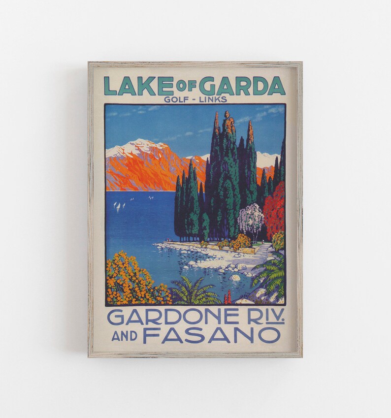 May include: Framed vintage travel poster for Lake Garda, Italy. The artwork features a scenic view of the lake with mountains, trees, and the text "LAKE OF GARDA" and "GARDONE RIV. AND FASANO". The poster has a distressed, aged look.