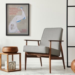 Op de afbeelding: Een moderne woonkamersc&egrave;ne met een ingelijste kunstprint, een stoel met hondenpoottjes en een houten bijzettafel. De kunstprint heeft een pauwenillustratie met tekst. Een houten kom en decoratieve dozen staan op de bijzettafel.