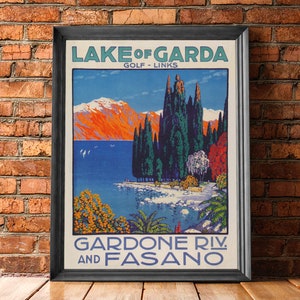May include: Framed vintage travel poster for Lake Garda, Italy, featuring a scenic landscape with mountains, water, and trees. The poster has text that reads "Lake of Garda" and "Gardone Riv. and Fasano."