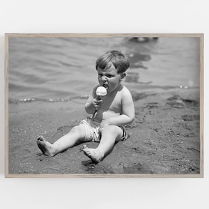 May include: A young boy sits on a sandy beach, wearing a swimsuit and eating an ice cream cone. He is looking at the camera with a serious expression.