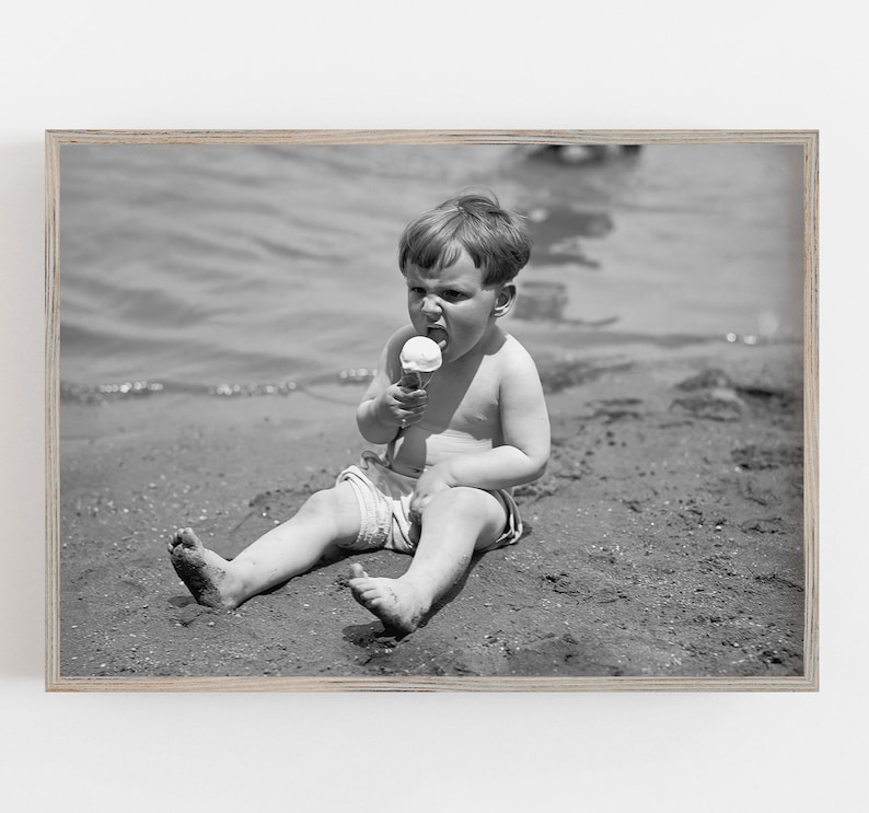 May include: A young boy sits on a sandy beach, wearing a swimsuit and eating an ice cream cone. He is looking at the camera with a serious expression.