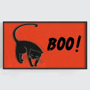May include: A framed Halloween art print featuring a black cat silhouette with the word "BOO!" in bold black letters on a textured orange background. The cat is depicted with its tail arched.