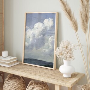 May include: A framed painting of a large, fluffy white cloud against a blue sky. The painting is hanging on a wall above a wooden bench with a woven seat.