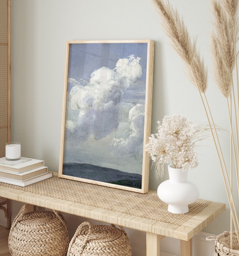 May include: A framed painting of a large, fluffy white cloud against a blue sky. The painting is hanging on a wall above a wooden bench with a woven seat.