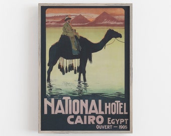 May include: Vintage travel poster for the National Hotel in Cairo, Egypt, featuring a person riding a camel in water, with pyramids in the background. The poster is framed and includes the text "OUVERT - 1905".