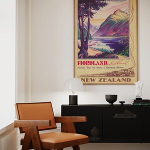Puede incluir: Un p&oacute;ster de viaje vintage enmarcado para Fiordland, Southland, Nueva Zelanda, con un paisaje de monta&ntilde;as, un lago y un tren. Una silla moderna de mediados de siglo con estructura de madera marr&oacute;n y asiento de cuero marr&oacute;n claro est&aacute; en primer plano.