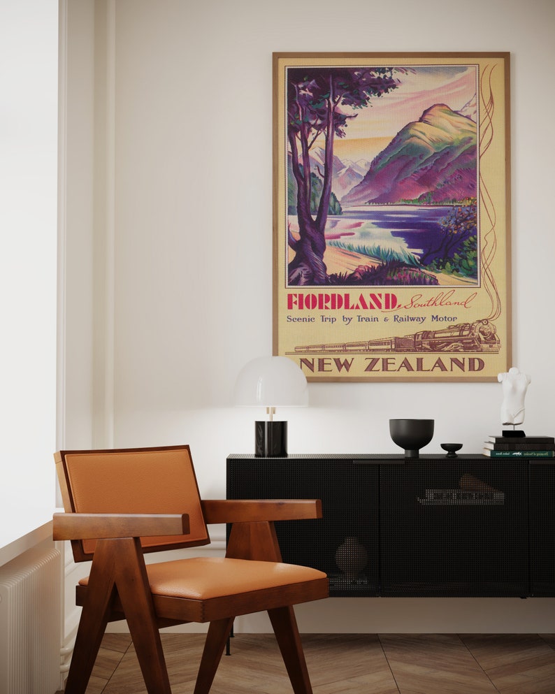Puede incluir: Un p&oacute;ster de viaje vintage enmarcado para Fiordland, Southland, Nueva Zelanda, con un paisaje de monta&ntilde;as, un lago y un tren. Una silla moderna de mediados de siglo con estructura de madera marr&oacute;n y asiento de cuero marr&oacute;n claro est&aacute; en primer plano.