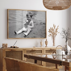 May include: Black and white photograph of a young boy sitting on a beach, eating an ice cream cone. The photo is framed in a wooden frame and hanging on a white wall.