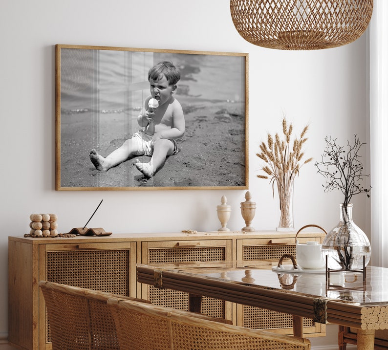 May include: Black and white photograph of a young boy sitting on a beach, eating an ice cream cone. The photo is framed in a wooden frame and hanging on a white wall.