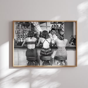 Op de afbeelding: Zwart-witfoto van vier mensen die aan een bar zitten. Drie vrouwen dragen badpakken en een man draagt een overhemd en een stropdas. De vrouwen zitten op krukken en de man staat. Er staan flessen op planken achter hen.