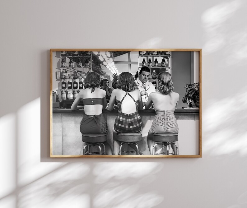 Op de afbeelding: Zwart-witfoto van vier mensen die aan een bar zitten. Drie vrouwen dragen badpakken en een man draagt een overhemd en een stropdas. De vrouwen zitten op krukken en de man staat. Er staan flessen op planken achter hen.