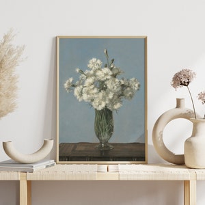 以下が含まれることがあります： 透明なガラスの花瓶に白い花が活けられた静物画の額入りアートプリント。花瓶は、明るい青色の背景に対して、濃い木製の表面に置かれています。アートワークは、装飾的な花瓶のある明るい木製のベンチに展示されています。