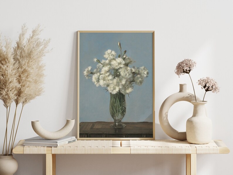 以下が含まれることがあります： 透明なガラスの花瓶に白い花が活けられた静物画の額入りアートプリント。花瓶は、明るい青色の背景に対して、濃い木製の表面に置かれています。アートワークは、装飾的な花瓶のある明るい木製のベンチに展示されています。