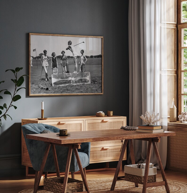 May include: A black and white photograph of five women playing golf on a green course. The women are wearing 1920s clothing and are standing on a block of ice. The photograph is framed in a wooden frame and is hanging on a dark gray wall.