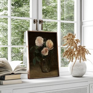 May include: A framed floral still life painting featuring three light pink roses in a glass vase. The painting is set on a white windowsill with a view of green foliage through a window.