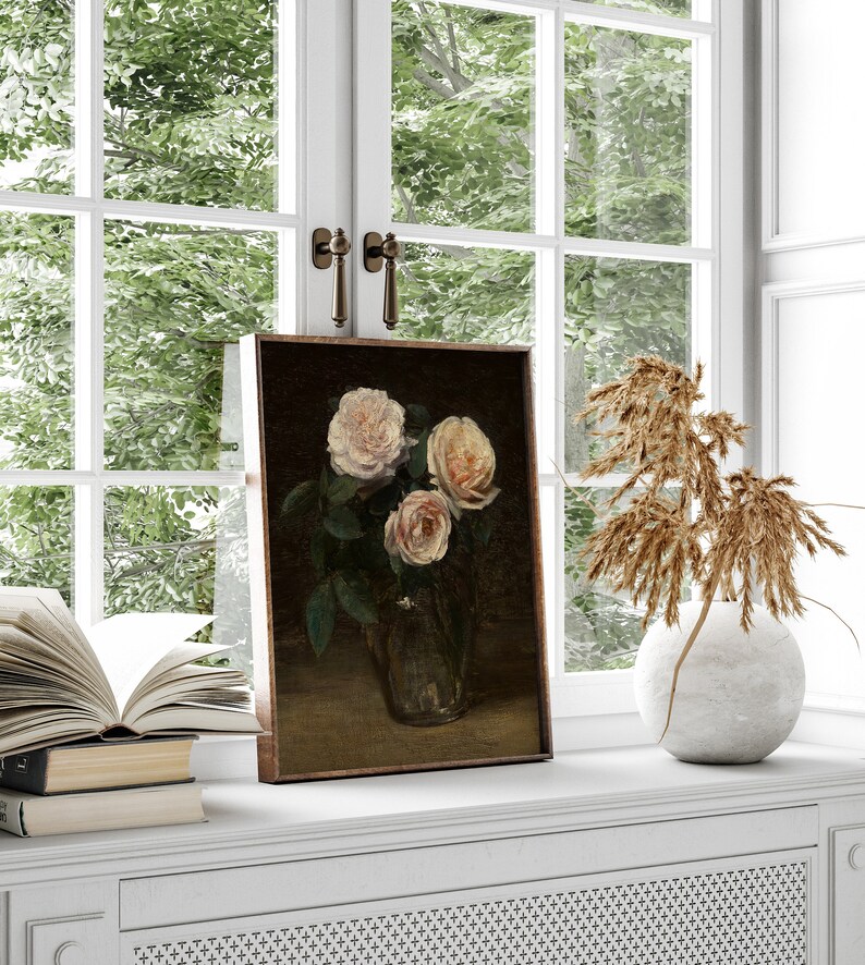 May include: A framed floral still life painting featuring three light pink roses in a glass vase. The painting is set on a white windowsill with a view of green foliage through a window.