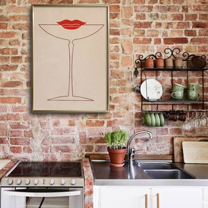 May include: A framed wall art print featuring a minimalist red line drawing of a martini glass with a pair of red lips positioned above the rim, set against a light beige background, displayed in a gold-coloured frame on a brick wall.