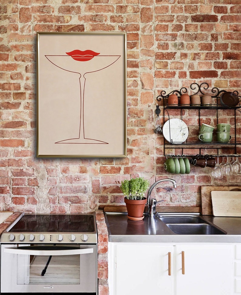 May include: A framed wall art print featuring a minimalist red line drawing of a martini glass with a pair of red lips positioned above the rim, set against a light beige background, displayed in a gold-coloured frame on a brick wall.