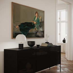 May include: A black metal cabinet with a wire mesh front, a white lamp with a white shade, a black bowl, and a stack of books on top. A framed painting of a woman in a green dress reading a book hangs on the wall above the cabinet.