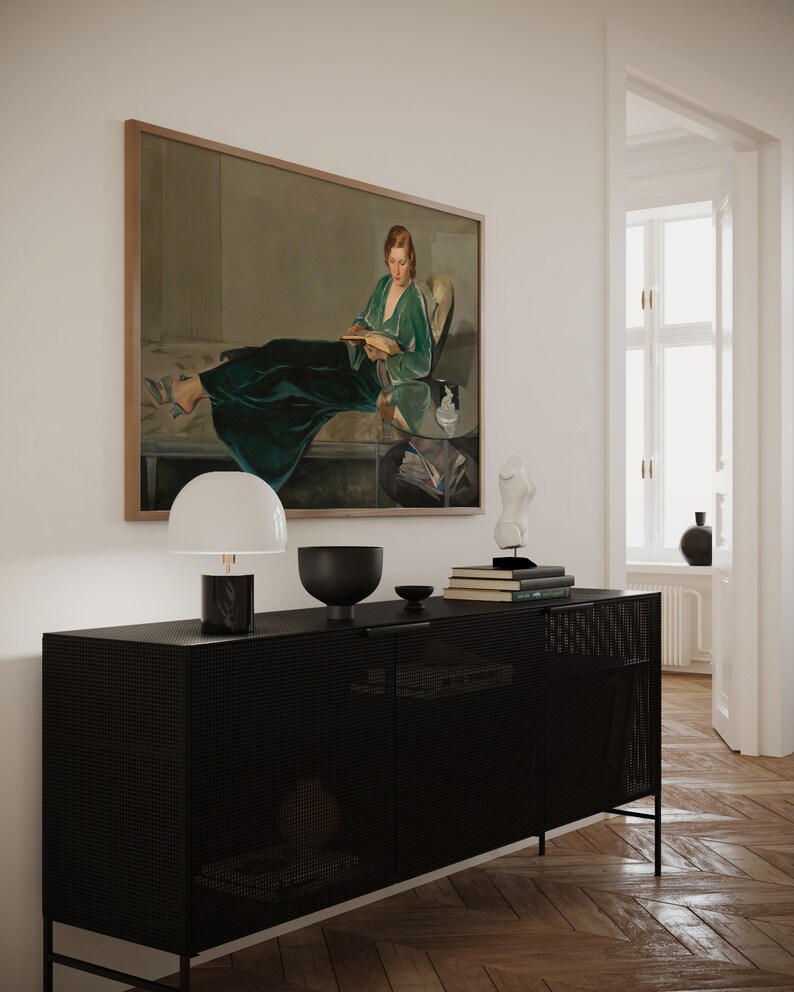 May include: A black metal cabinet with a wire mesh front, a white lamp with a white shade, a black bowl, and a stack of books on top. A framed painting of a woman in a green dress reading a book hangs on the wall above the cabinet.