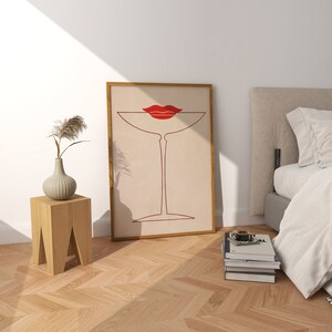 May include: A framed wall art print featuring a minimalist line drawing of a martini glass in dark red, topped with a pair of bright red lips. The artwork is set against a light beige background and enclosed in a simple wooden frame.