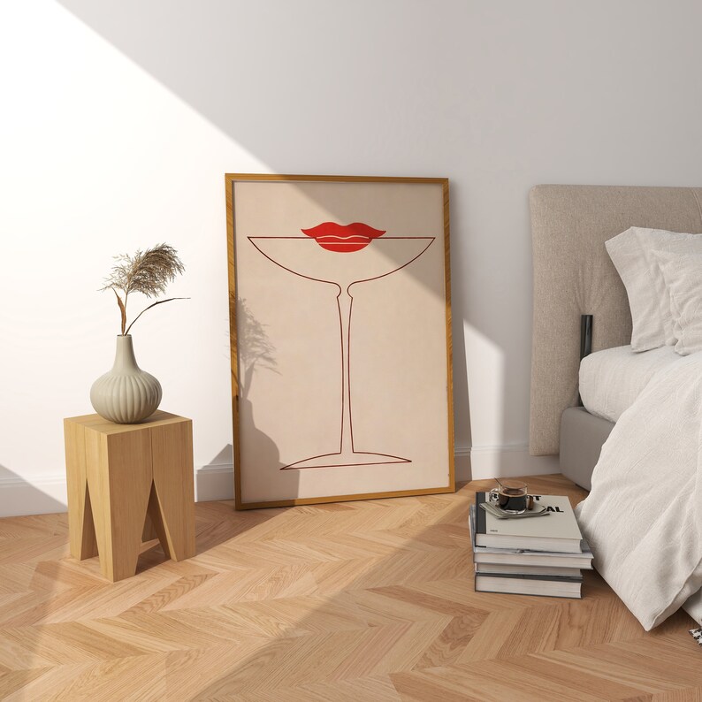 May include: A framed wall art print featuring a minimalist line drawing of a martini glass in dark red, topped with a pair of bright red lips. The artwork is set against a light beige background and enclosed in a simple wooden frame.