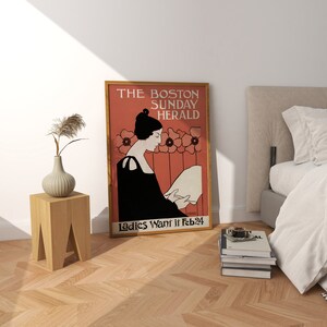 May include: A framed vintage poster with the text "THE BOSTON SUNDAY HERALD" and "Ladies Want it Feb 24". The poster features a woman in black attire reading, set against a red background with floral accents. A wooden side table and vase are in the foreground.