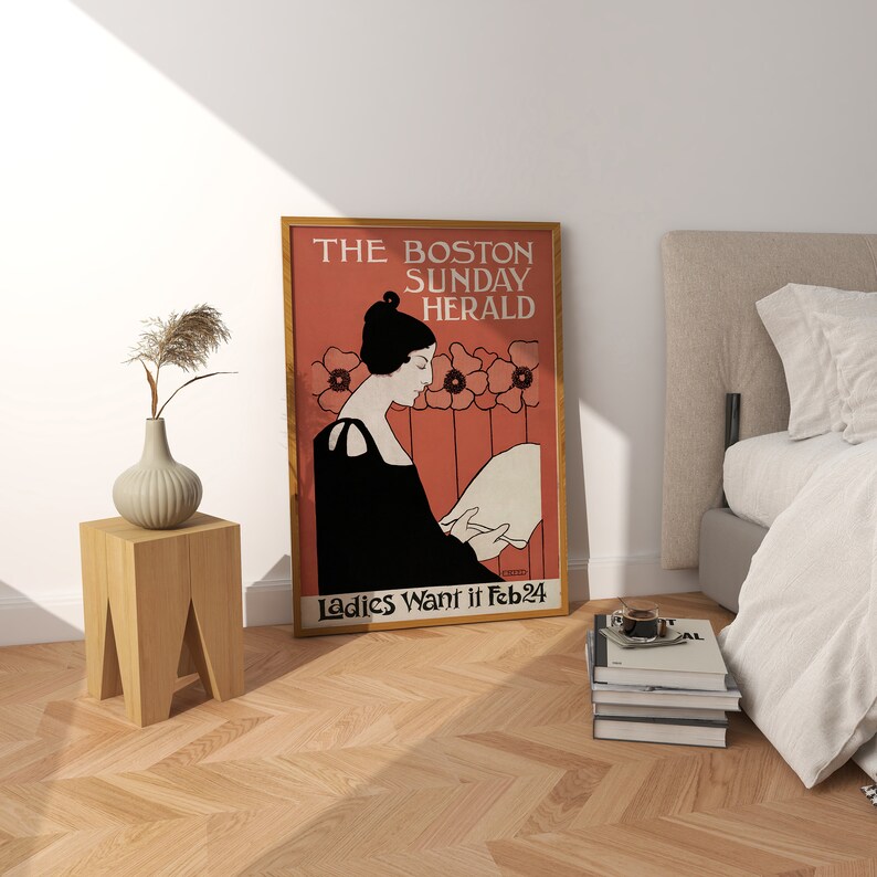 May include: A framed vintage poster with the text "THE BOSTON SUNDAY HERALD" and "Ladies Want it Feb 24". The poster features a woman in black attire reading, set against a red background with floral accents. A wooden side table and vase are in the foreground.
