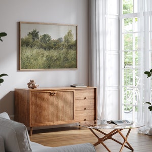 May include: A landscape art print in a wooden frame, showing a field of green grass and trees. The artwork is above a wooden cabinet with a woven door and three drawers. A glass-topped coffee table and white curtains are also visible.