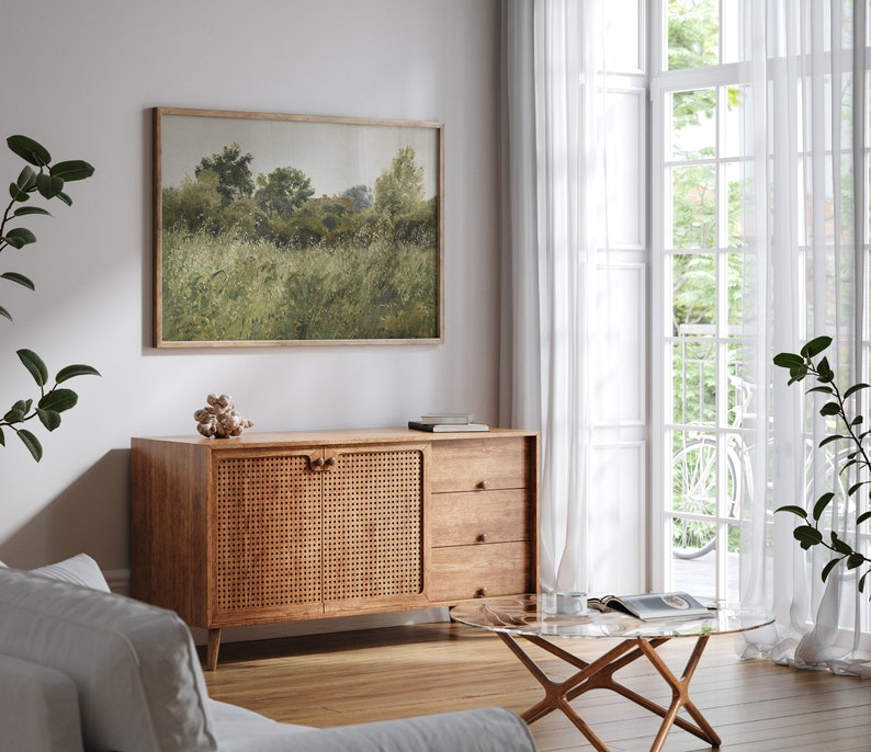 May include: A landscape art print in a wooden frame, showing a field of green grass and trees. The artwork is above a wooden cabinet with a woven door and three drawers. A glass-topped coffee table and white curtains are also visible.