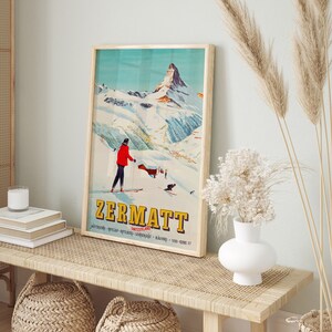Puede incluir: Un cartel de viaje vintage con un dise&ntilde;o retro que presenta a un esquiador en una ladera de monta&ntilde;a en Zermatt, Suiza. El cartel tiene un esquema de color azul, blanco y rojo y presenta el texto "ZERMATT" en letras amarillas.