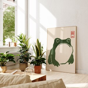 May include: A minimalist art print featuring a green frog with a white background. The print has a red square with Japanese characters in the top right corner.