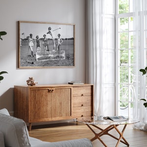 May include: A black and white photograph of four people playing golf. The photo is framed and hanging on a white wall above a wooden cabinet with a woven rattan door. The cabinet has two drawers below the door.