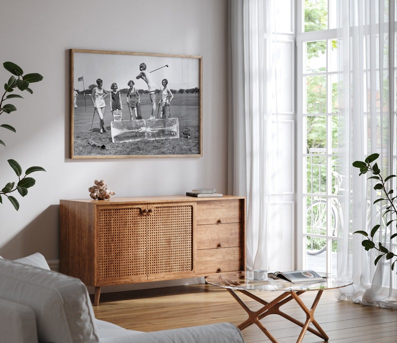 May include: A black and white photograph of four people playing golf. The photo is framed and hanging on a white wall above a wooden cabinet with a woven rattan door. The cabinet has two drawers below the door.