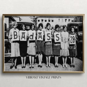 May include: Black and white photograph of seven women holding signs that spell out "BADASS" and "28". The women are wearing flapper-style dresses and are standing in front of a vintage car.
