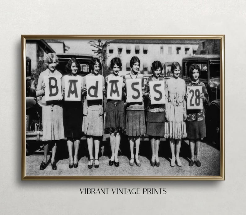 May include: Black and white photograph of seven women holding signs that spell out "BADASS" and "28". The women are wearing flapper-style dresses and are standing in front of a vintage car.