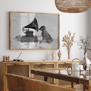 May include: A black and white photograph of a dog listening to a vintage gramophone. The dog is sitting on a snowy surface and the gramophone is on a wooden box. The photograph is framed and hanging on a white wall.