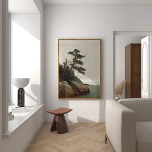 May include: A framed landscape painting of a tree on a rocky cliff overlooking a body of water. The painting is in a wooden frame and is hanging on a white wall.