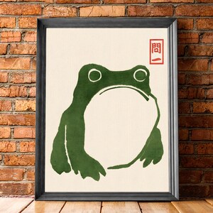 May include: A framed print of a green frog silhouette with a red Japanese character "開" in the top right corner. The background is a cream color.