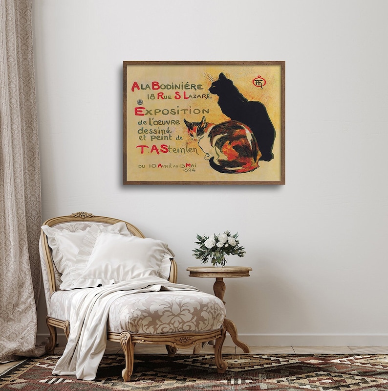 May include: A framed vintage poster featuring two cats, one black and one calico, against a tan background. The poster includes French text, advertising an exhibition of artwork by T.A. Steinlen. The poster is displayed on a white wall.