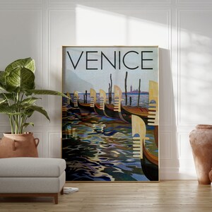 May include: A framed art print featuring the word "VENICE" in large black letters above a depiction of gondolas on the water. The artwork has a vintage travel poster style with a gold frame. The scene includes gondolas, water reflections, and a distant cityscape.