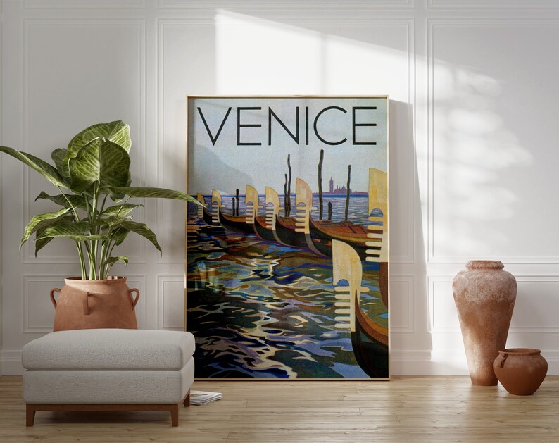 May include: A framed art print featuring the word "VENICE" in large black letters above a depiction of gondolas on the water. The artwork has a vintage travel poster style with a gold frame. The scene includes gondolas, water reflections, and a distant cityscape.