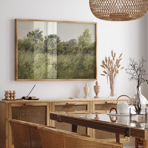May include: A framed landscape painting in a wooden frame hangs above a wooden cabinet. The painting shows a field of tall grass and trees in green and grey tones. A woven pendant light hangs from the ceiling. The room is decorated with natural elements.